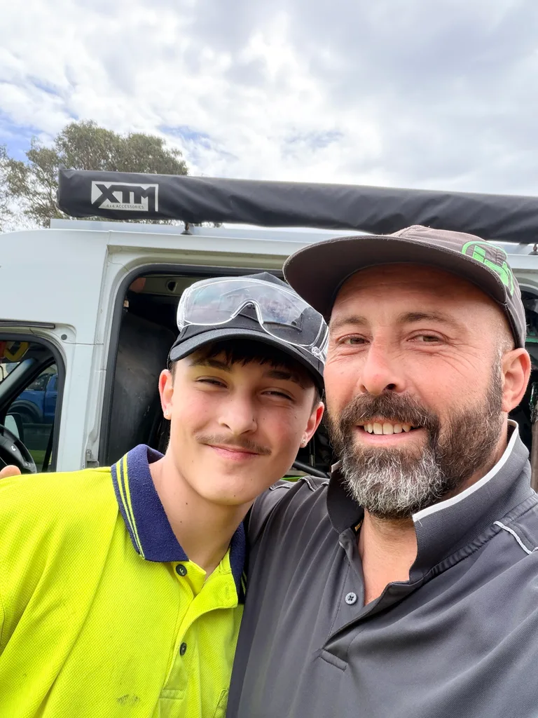 James and his son Lucas smiling in front of their white work van, Lucas in a hi-vis shirt and James in a cap