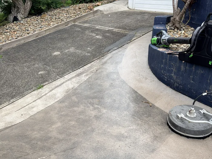 Pressure washing a concrete driveway in progress: the cleaned half is pale, the uncleaned half still grey, with the surface cleaner sitting on the concrete
