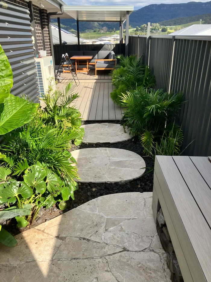 Round stone stepping pads set in black pebbles through a tidy tropical garden, with a timber deck and the escarpment behind