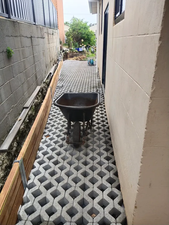 Narrow side walkway laid with grey grass-grid pavers between a block retaining wall and a rendered house wall, with a black wheelbarrow on the path